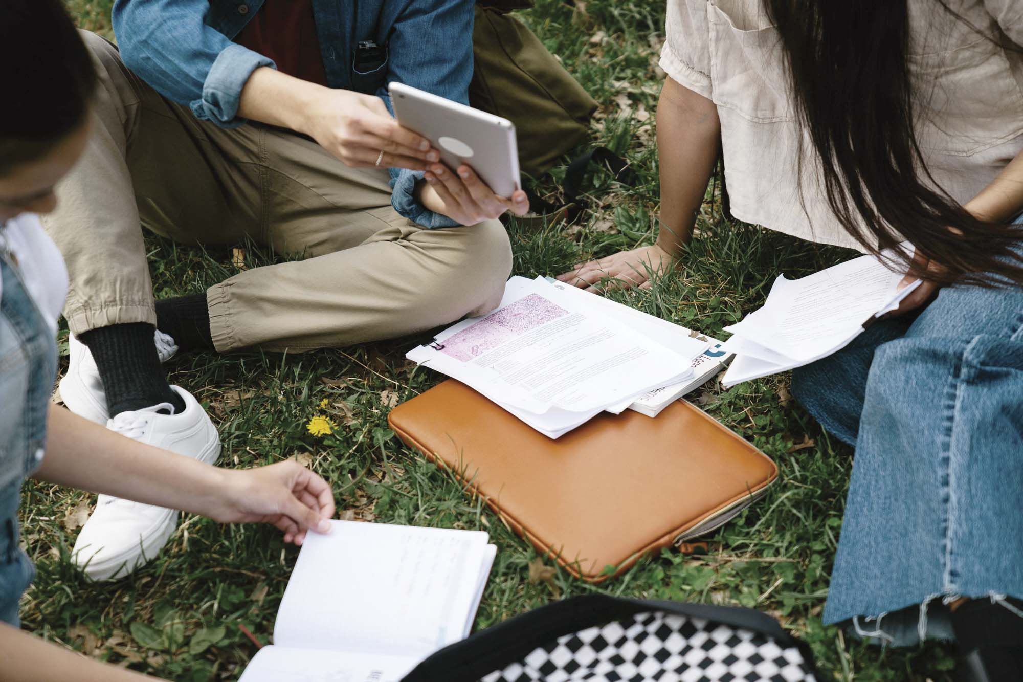 Close-up overhead view of students sitting in a circle on the lawn with study papers, a tablet, and a leather folder