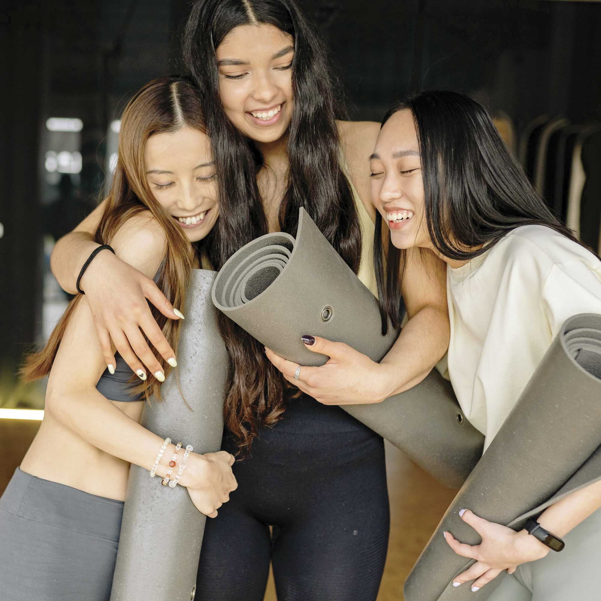 Three young women laughing and hugging in a fitness studio while holding rolled-up grey yoga mats